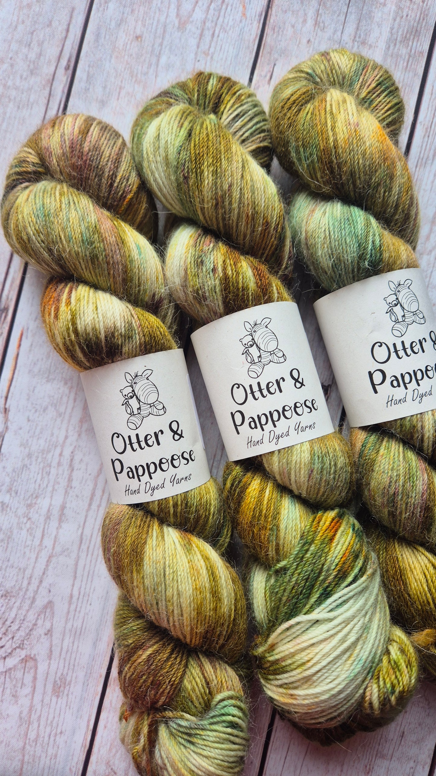 A row of three hand-dyed yarn skeins from Otter & Pappoose, showcasing a variegated and speckled colorway of natural earthy tones including deep olive green, golden yellow, orange, warm brown, and hints of teal and orange, lying on a light wooden background.