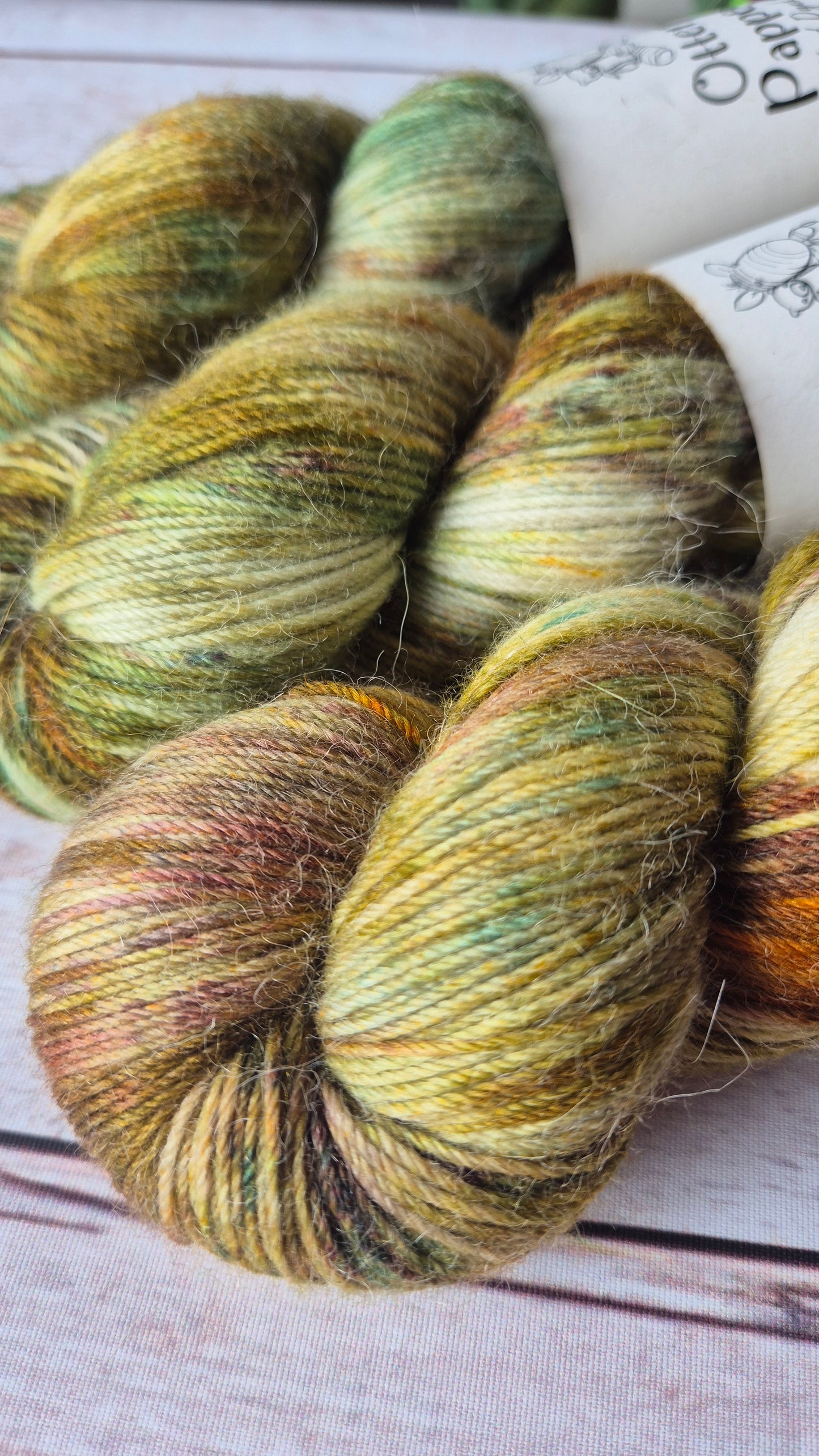 A close-up of skeins by Otter & Pappoose hand-dyed yarn, displaying a rich, mottled colour palette of deep olive green, warm golden yellow, and various shades of brown, with hints of blue-green and orange rust, on a light wooden background.
