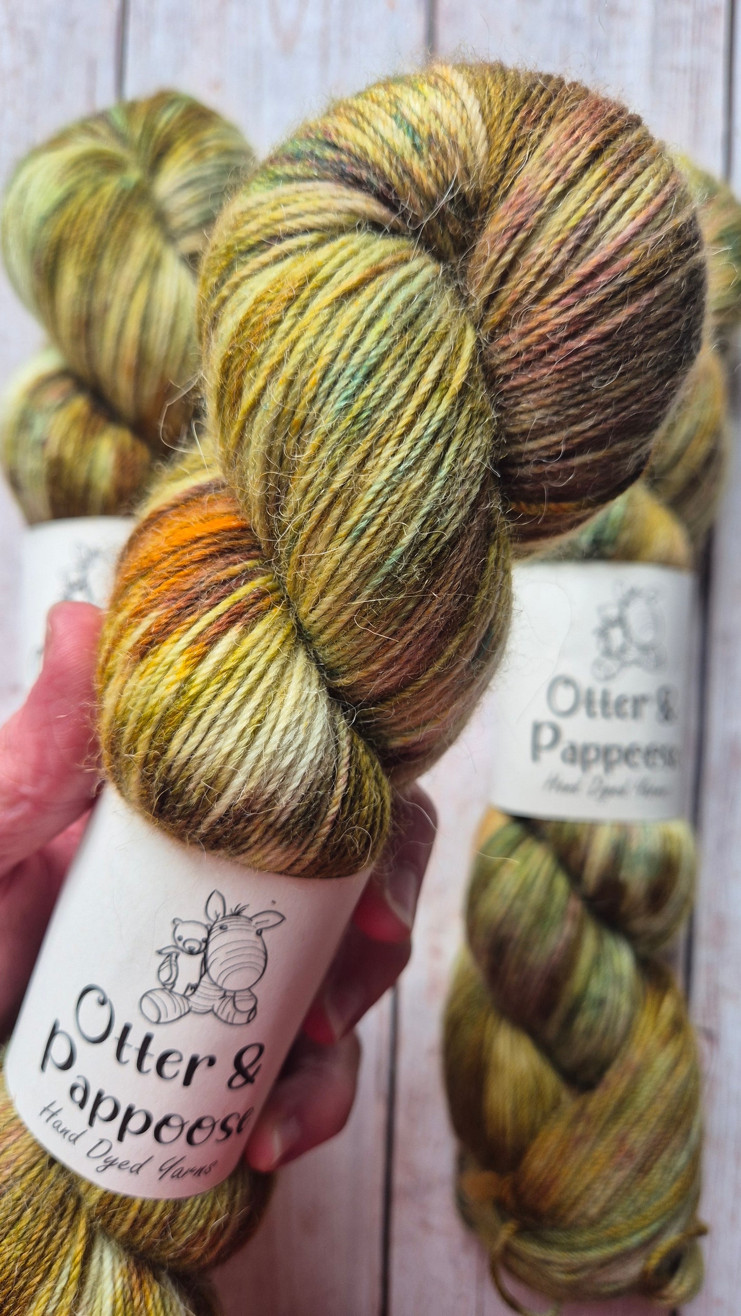 Close-up of a skein of Otter & Pappoose hand-dyed yarn, showcasing mottled tones of olive green, rusty orange, deep moss, and hints of a reddish-brown and teal speckles, held against a wooden background.