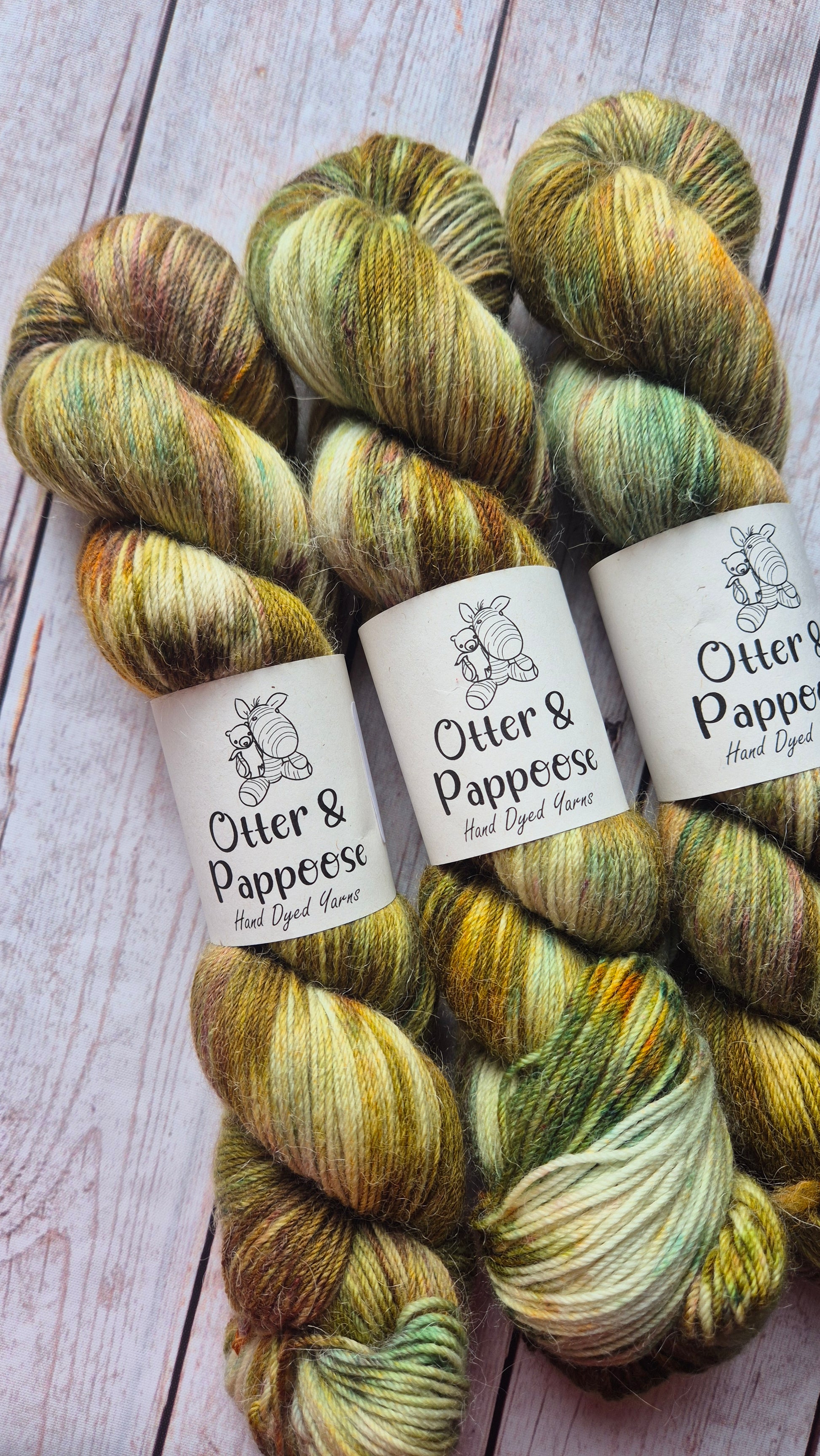 A row of three hand-dyed yarn skeins from Otter & Pappoose, showcasing a variegated and speckled colorway of natural earthy tones including deep olive green, golden yellow, orange, warm brown, and hints of teal and orange, lying on a light wooden background.